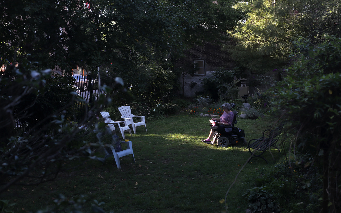A dark color photograph of a woman in a wheelchair reading in a garden.